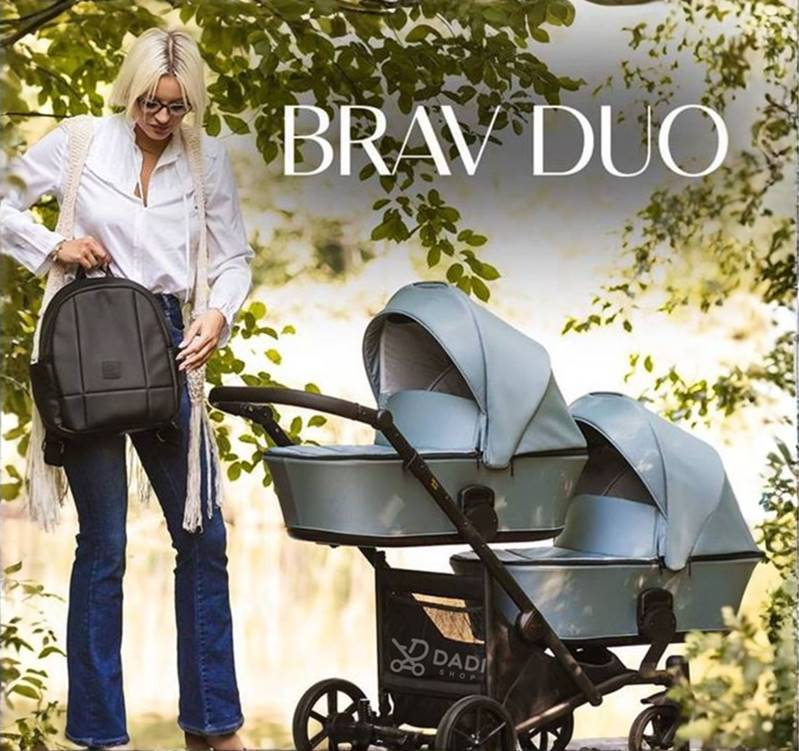 Stylowy i lekki wózek dla bliźniąt Kunert Brav Duo w błękitnym kolorze. Kobieta z plecakiem prezentuje model na spacerze wśród zieleni.